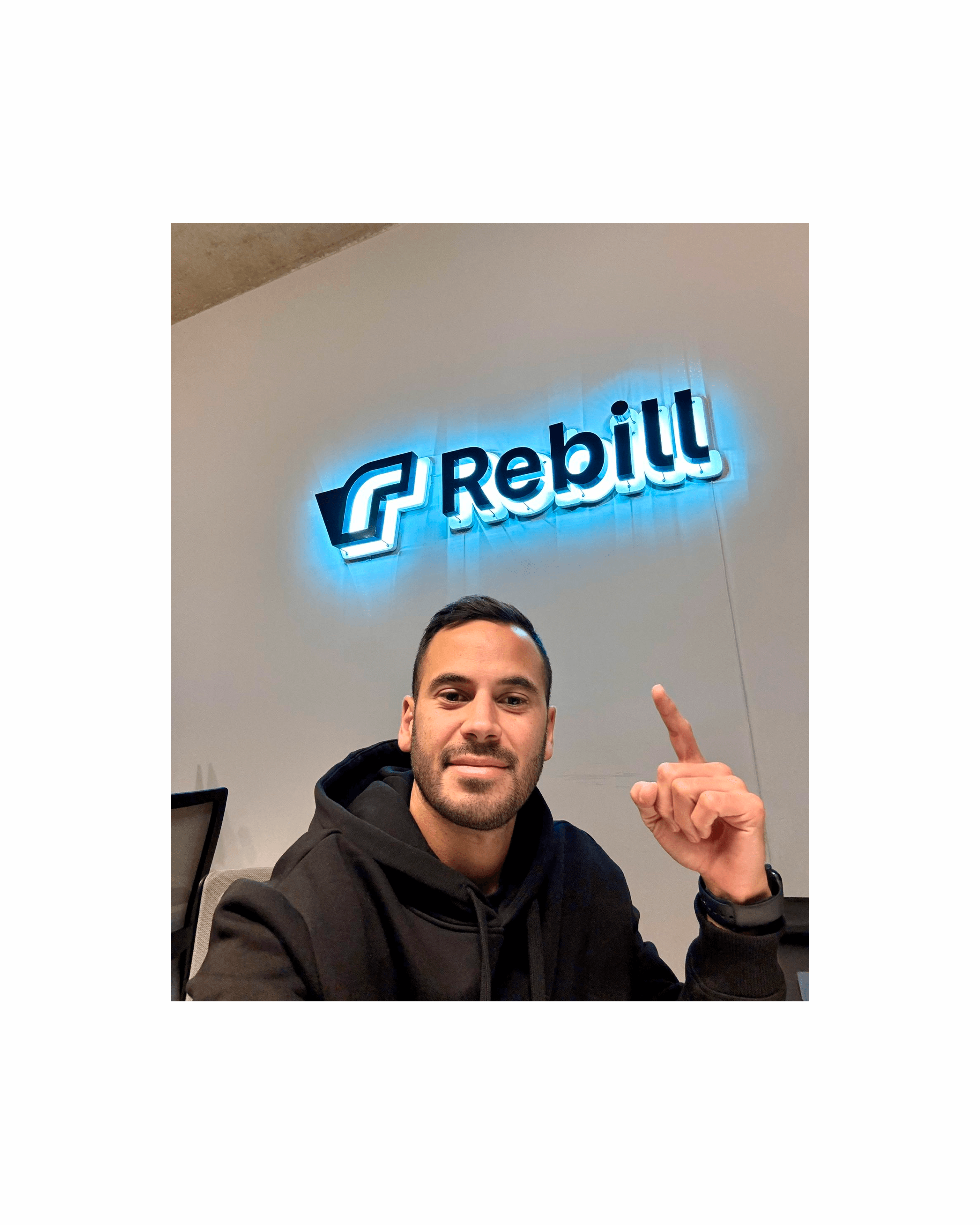 Photo of Rebill’s illuminated logo installed in the office—founder Nahuel Candia pointing to the new sign alongside the modern workspace—showcasing how Kundo Studio’s refreshed visual identity brings the Stripe-grade LATAM payments brand into its physical environment.
