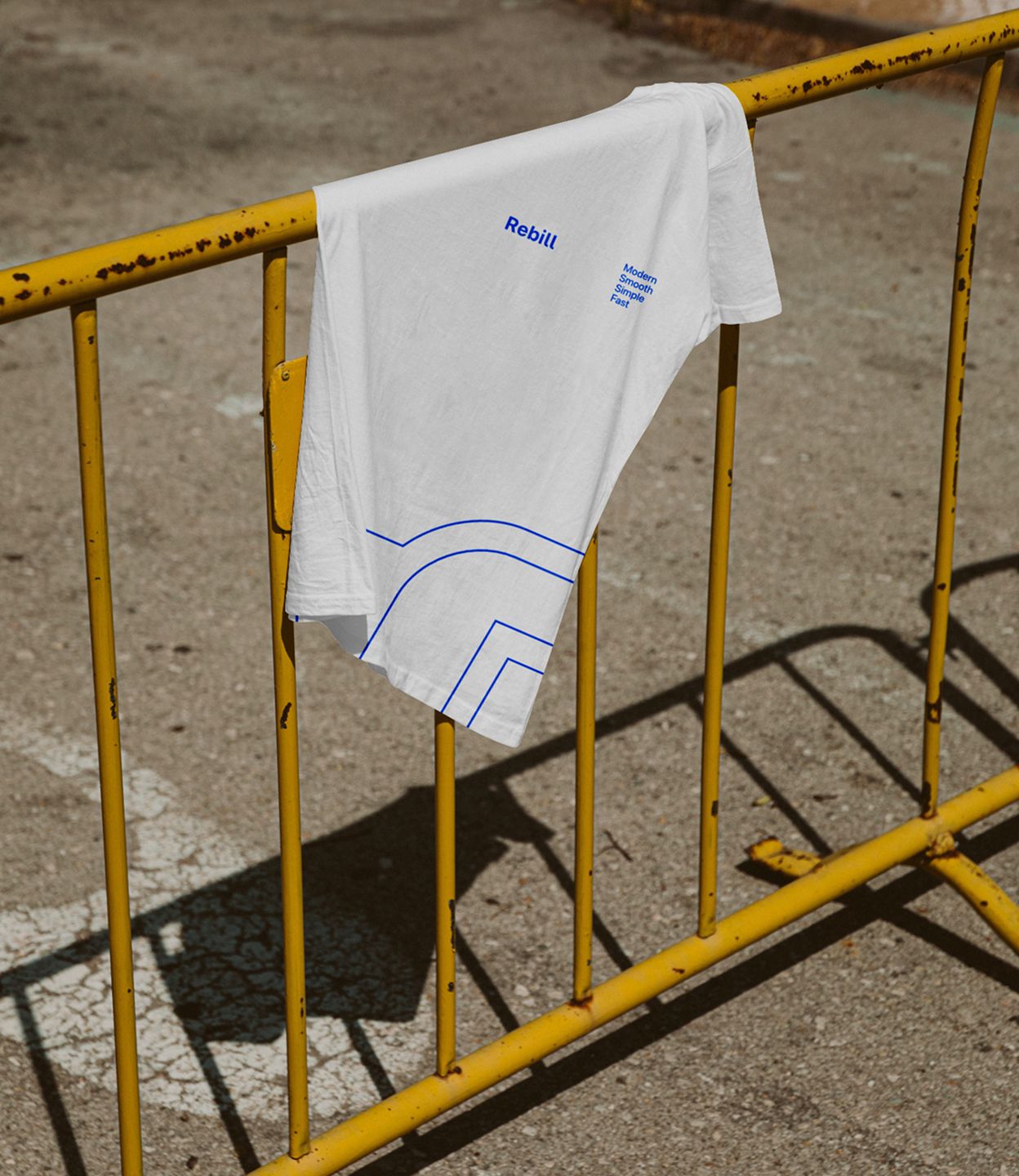 White t-shirt with oversized blue Rebill logo line graphic draped over a yellow street barrier, showcasing Kundo Studio’s confident, minimal visual identity for the LATAM payments infrastructure brand.