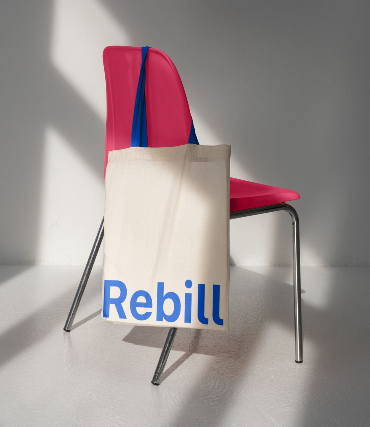 Canvas tote bag with bold blue Rebill wordmark hanging on a pink chair, showing how Kundo Studio’s refreshed payments infrastructure brand extends into simple, confident physical collateral for the LATAM market.